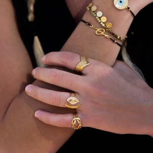 May include: Gold-toned jewelry set featuring rings and bracelets. The rings include a chevron design, an eye motif, and a knot style. Bracelets with gold disc and snake charms are also visible. The jewelry is worn on a person's arm.