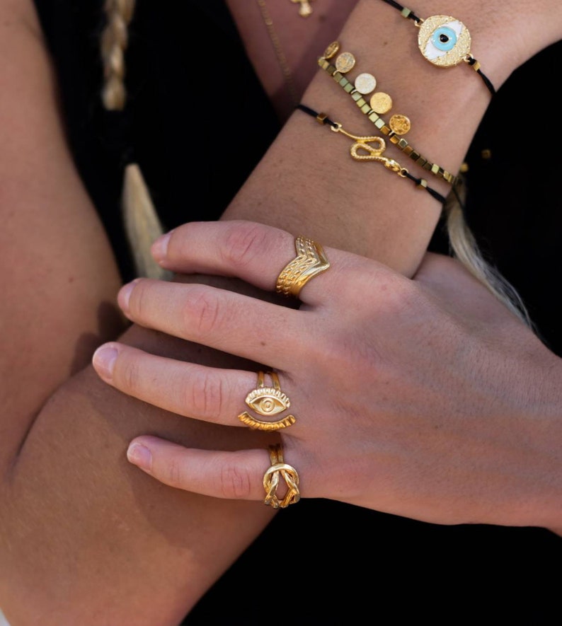 May include: Gold-toned jewelry set featuring rings and bracelets. The rings include a chevron design, an eye motif, and a knot style. Bracelets with gold disc and snake charms are also visible. The jewelry is worn on a person's arm.