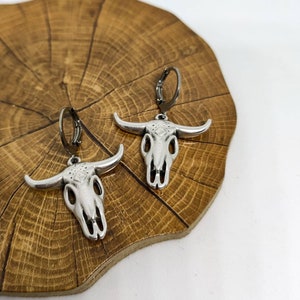 May include: Silver-toned bull skull earrings with black leverback closures. The earrings feature detailed skull designs with prominent horns. The jewelry is displayed on a wooden surface with a natural grain pattern, against a white background.