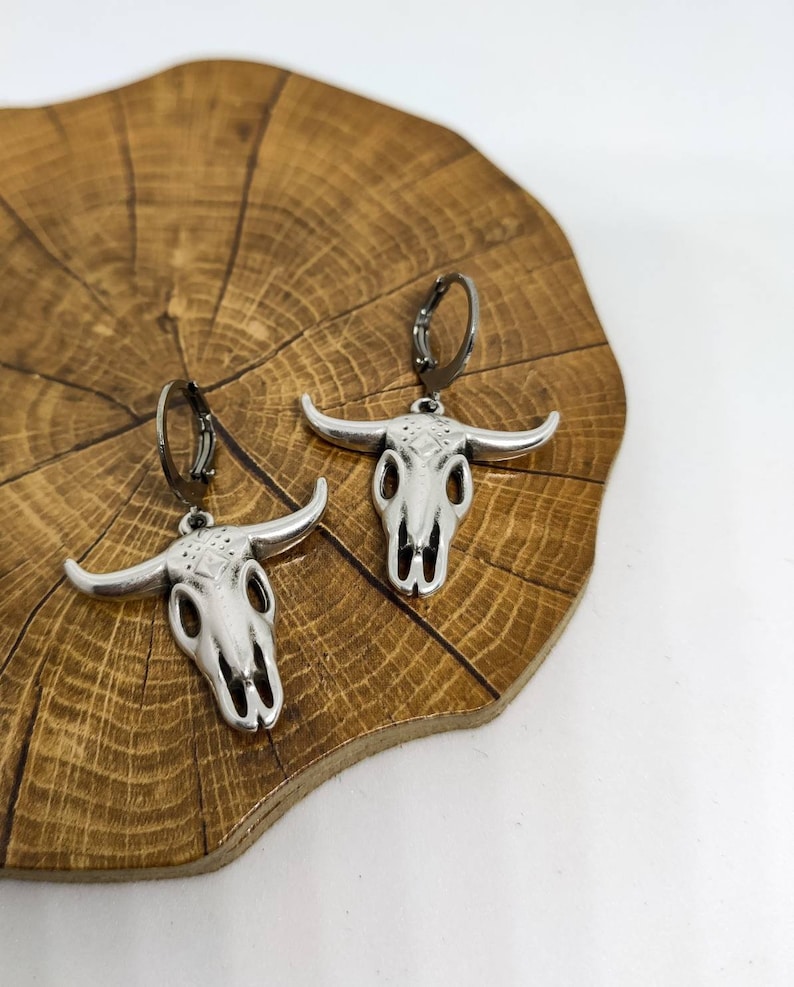 May include: Silver-toned bull skull earrings with black leverback closures. The earrings feature detailed skull designs with prominent horns. The jewelry is displayed on a wooden surface with a natural grain pattern, against a white background.