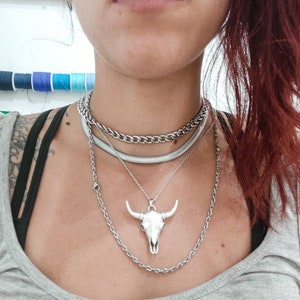 May include: A silver chain necklace with a silver bull skull pendant. The necklace is layered with two other silver chains.