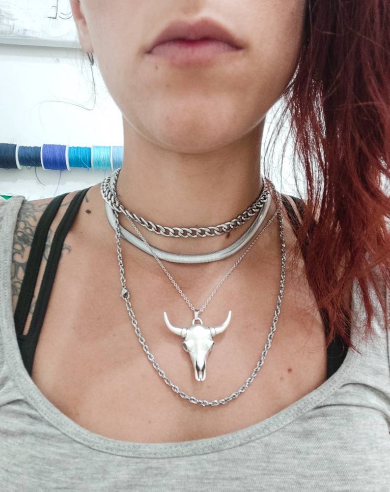 May include: A silver chain necklace with a silver bull skull pendant. The necklace is layered with two other silver chains.