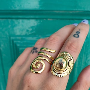 May include: Two gold rings are shown on a hand. One ring has a spiral design, the other has a large, oval face with a textured border and a central sphere. The rings are set against a teal background.