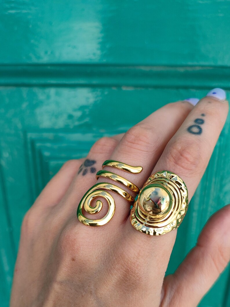 May include: Two gold rings are shown on a hand. One ring has a spiral design, the other has a large, oval face with a textured border and a central sphere. The rings are set against a teal background.