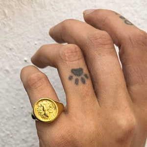 Op de afbeelding: Een goudkleurige zegelring met een gedetailleerd bijenontwerp. De ring heeft een ronde voorkant en een dikke band. De ring wordt gedragen op een vinger met een pootafdruk-tattoo.