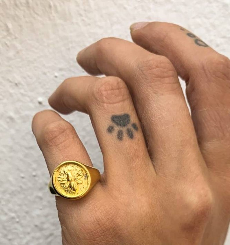 Op de afbeelding: Een goudkleurige zegelring met een gedetailleerd bijenontwerp. De ring heeft een ronde voorkant en een dikke band. De ring wordt gedragen op een vinger met een pootafdruk-tattoo.