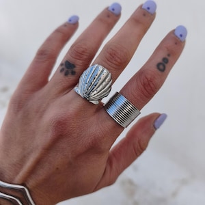 May include: A hand displays two silver rings: a seashell design and a wide band with horizontal lines. The hand also has a silver bracelet and light purple nail polish. The background is a blurred white.