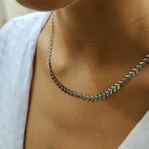 Op de afbeelding: Een zilverkleurige ketting met een chevron-ontwerp. De ketting is gemaakt van kleine, met elkaar verbonden pijlvormige stukjes. De ketting wordt gedragen over een wit overhemd.