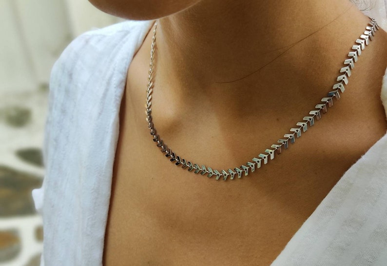 Op de afbeelding: Een zilverkleurige ketting met een chevron-ontwerp. De ketting is gemaakt van kleine, met elkaar verbonden pijlvormige stukjes. De ketting wordt gedragen over een wit overhemd.