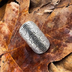 Könnte beinhalten: Ein silberfarbener, zylindrischer Ring mit aufwendigen Blumen- und Blattmustern. Der Ring ist sehr detailliert und scheint aus Metall zu sein. Der Ring liegt auf einem braunen Blatt.