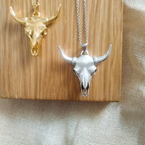 May include: Two silver and gold bull skull necklaces on a wooden surface. The necklaces are both on chains and have a detailed skull design.