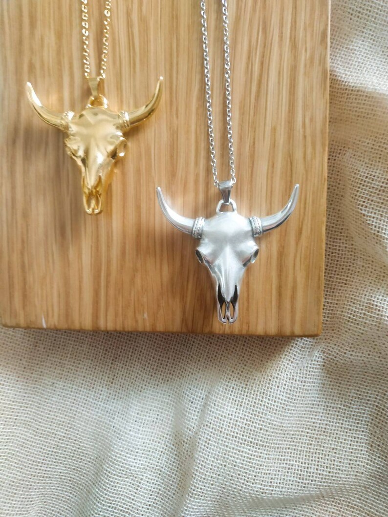 May include: Two silver and gold bull skull necklaces on a wooden surface. The necklaces are both on chains and have a detailed skull design.