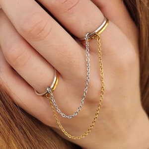 May include: A two-finger ring with a gold and silver chain. The ring has a gold band and a silver band, linked by a delicate chain. The chain is made of small, linked segments, creating a stylish and unique accessory.