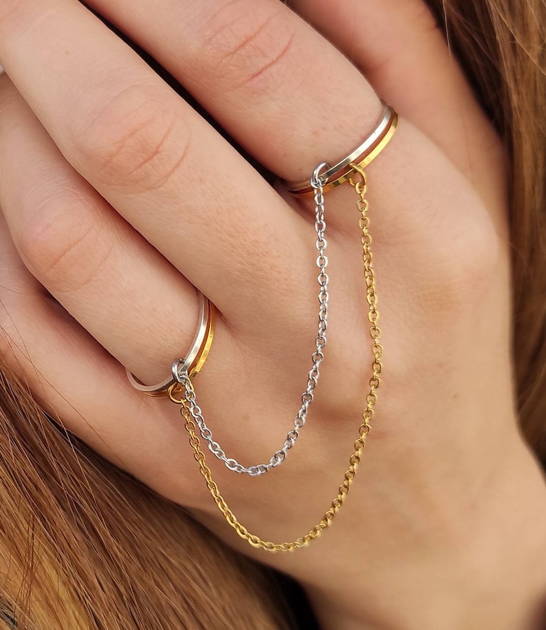 May include: A two-finger ring with a gold and silver chain. The ring has a gold band and a silver band, linked by a delicate chain. The chain is made of small, linked segments, creating a stylish and unique accessory.