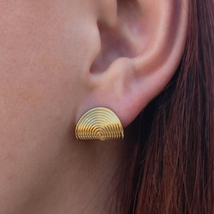Puede incluir: Un pendiente de botón dorado con forma de semicírculo y diseño en espiral. El pendiente se lleva en la oreja, mostrando sus intrincados detalles y acabado pulido. La joya añade un toque de elegancia.