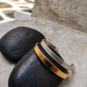 Pode incluir: Tr&ecirc;s pulseiras minimalistas de metal. Uma &eacute; dourada e as outras duas s&atilde;o prateadas. As pulseiras s&atilde;o exibidas sobre uma pedra preta e lisa, com um fundo desfocado. As pulseiras t&ecirc;m um design simples e elegante.