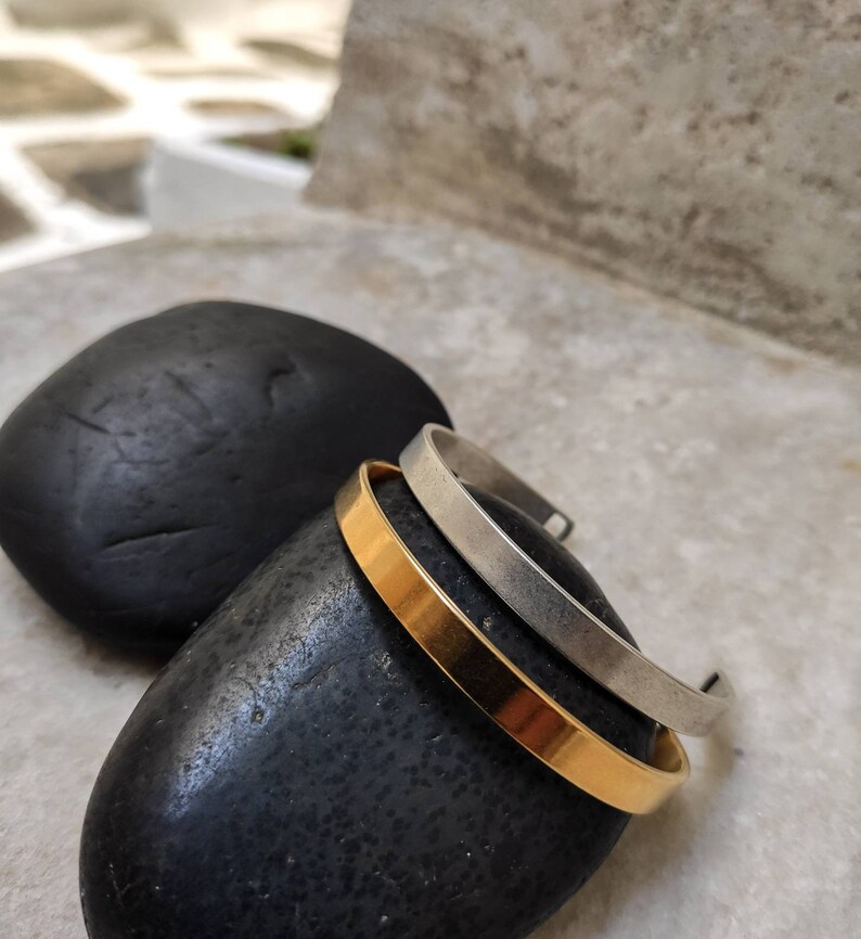 Pode incluir: Tr&ecirc;s pulseiras minimalistas de metal. Uma &eacute; dourada e as outras duas s&atilde;o prateadas. As pulseiras s&atilde;o exibidas sobre uma pedra preta e lisa, com um fundo desfocado. As pulseiras t&ecirc;m um design simples e elegante.