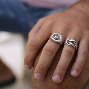 Pu&ograve; includere: Due anelli d'argento sulla mano di una persona. Un anello ha un design a occhio in rilievo, l'altro &egrave; un design a corda intrecciata.