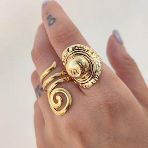 May include: A gold ring featuring a spiral design and a large, textured oval centrepiece. The ring wraps around the finger with a swirling band and a decorative, domed top.