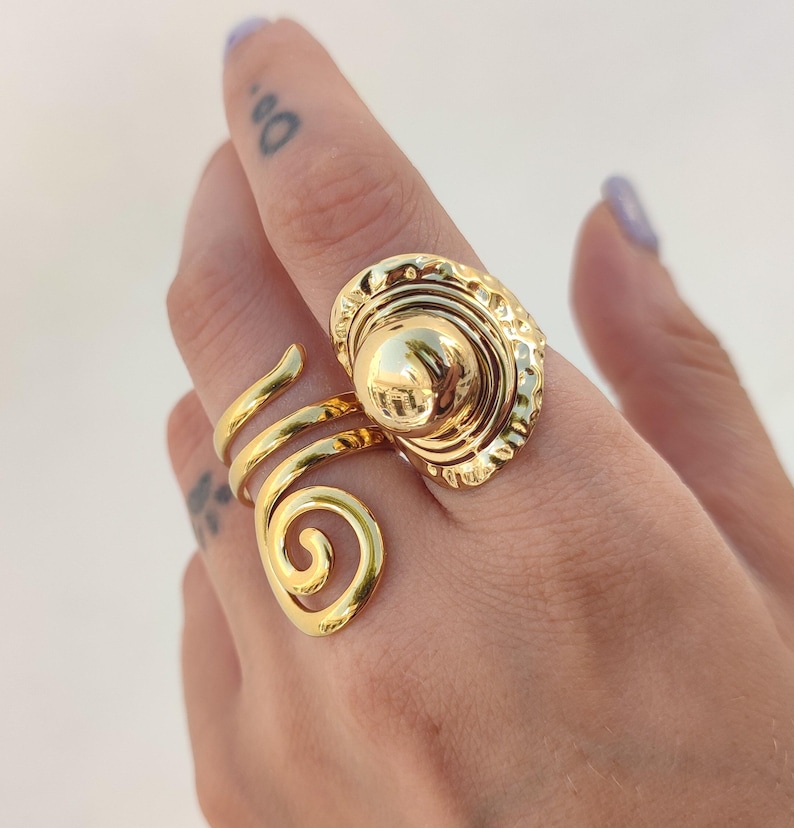 May include: A gold ring featuring a spiral design and a large, textured oval centrepiece. The ring wraps around the finger with a swirling band and a decorative, domed top.