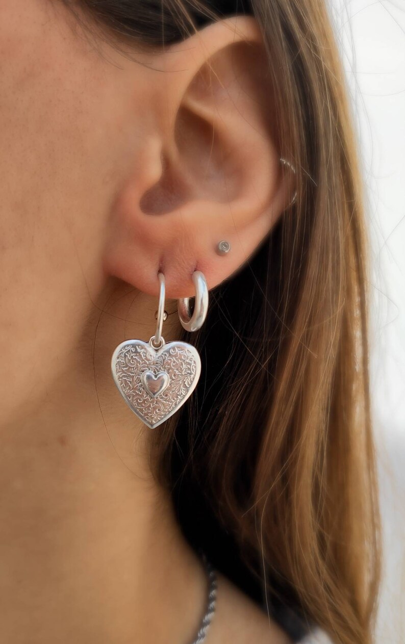 Puede incluir: Pendientes de aro plateados con forma de coraz&oacute;n. El colgante en forma de coraz&oacute;n presenta un intrincado dise&ntilde;o floral y un coraz&oacute;n m&aacute;s peque&ntilde;o en el centro. Tambi&eacute;n se ve un pendiente de aro plateado m&aacute;s peque&ntilde;o. Los pendientes se llevan en la oreja.