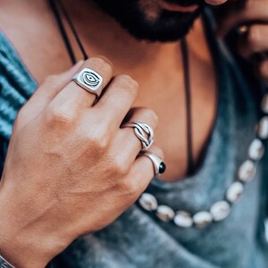 Pu&ograve; includere: Una persona indossa un anello d'argento con un designo a forma di occhio, un anello d'argento con un designo a forma di nodo e un anello d'argento con una pietra nera.