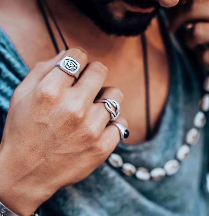 Pu&ograve; includere: Una persona indossa un anello d'argento con un designo a forma di occhio, un anello d'argento con un designo a forma di nodo e un anello d'argento con una pietra nera.