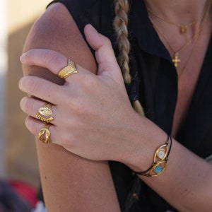 May include: A hand adorned with gold rings and a bracelet. The rings feature various designs, including an eye and a textured chevron shape. The bracelet has two gold-framed eye-shaped charms with white and turquoise centers.