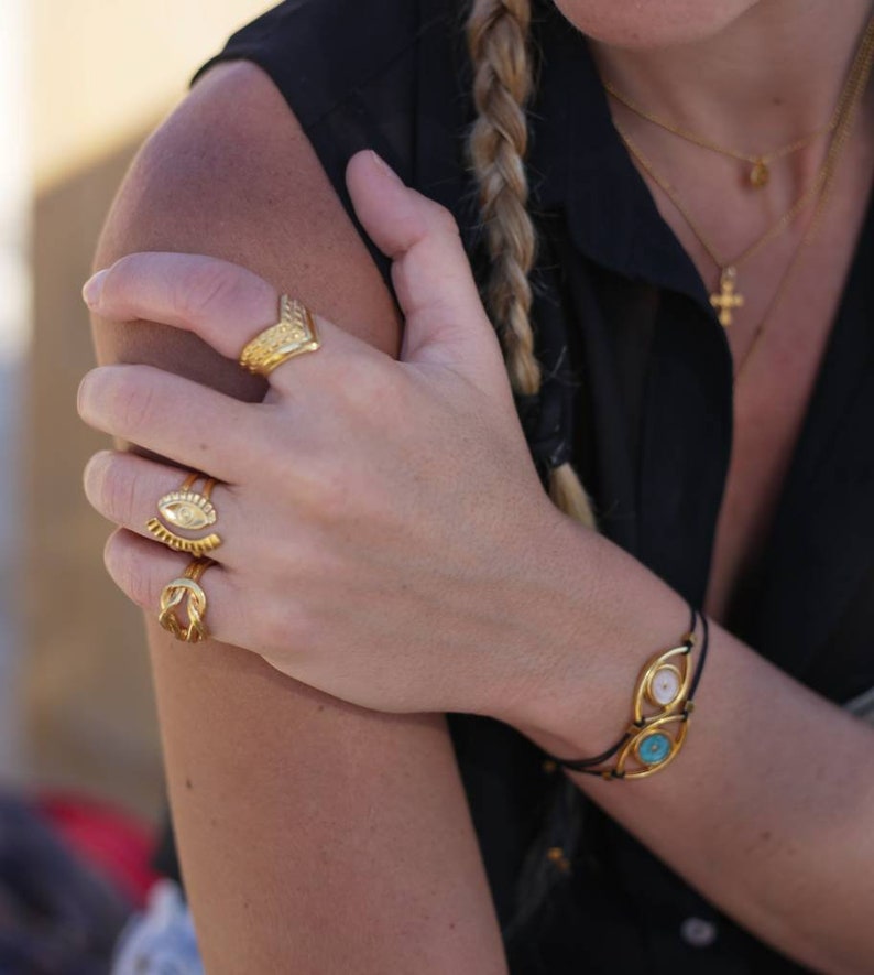 May include: A hand adorned with gold rings and a bracelet. The rings feature various designs, including an eye and a textured chevron shape. The bracelet has two gold-framed eye-shaped charms with white and turquoise centers.