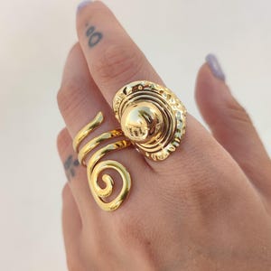 May include: A gold ring with a spiral design and a large, textured oval centerpiece. The ring wraps around the finger with a swirling band and features a raised, circular element within the oval.
