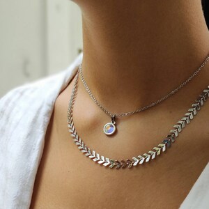 Op de afbeelding: Een zilveren ketting set met twee strengen. De bovenste ketting heeft een kleine ronde hanger met iriserende kleuren. De onderste ketting heeft een chevron ontwerp. De kettingen worden gedragen door een persoon die een wit overhemd draagt.