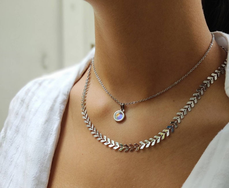 Op de afbeelding: Een zilveren ketting set met twee strengen. De bovenste ketting heeft een kleine ronde hanger met iriserende kleuren. De onderste ketting heeft een chevron ontwerp. De kettingen worden gedragen door een persoon die een wit overhemd draagt.