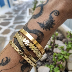 Pode incluir: Quatro pulseiras douradas de v&aacute;rios designs s&atilde;o empilhadas em um pulso com uma tatuagem. As pulseiras incluem uma faixa plana, uma faixa texturizada, um estilo de elos de corrente e um design tran&ccedil;ado. As pulseiras s&atilde;o exibidas em um fundo desfocado ao ar livre.