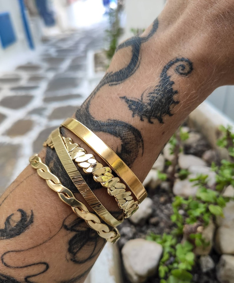 Pode incluir: Quatro pulseiras douradas de v&aacute;rios designs s&atilde;o empilhadas em um pulso com uma tatuagem. As pulseiras incluem uma faixa plana, uma faixa texturizada, um estilo de elos de corrente e um design tran&ccedil;ado. As pulseiras s&atilde;o exibidas em um fundo desfocado ao ar livre.