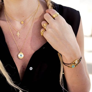 May include: A collection of gold-toned jewelry, including layered necklaces with a cross and an eye pendant, rings, and a bracelet with eye-shaped charms. The jewelry is worn with a black top. The bracelet has two eye-shaped charms with blue and pink stones.
