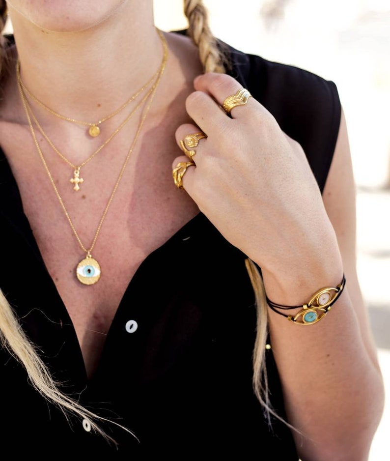May include: A collection of gold-toned jewelry, including layered necklaces with a cross and an eye pendant, rings, and a bracelet with eye-shaped charms. The jewelry is worn with a black top. The bracelet has two eye-shaped charms with blue and pink stones.