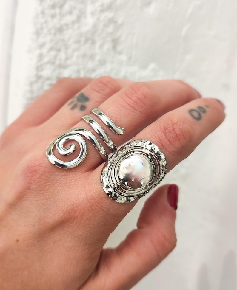 May include: Two silver rings on a finger. One ring is a spiral design and the other is a round ring with a hammered texture.