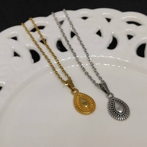 May include: Two necklaces with teardrop pendants. One necklace is gold-toned, the other is silver-toned. Both necklaces have delicate chains and are displayed on a white, decorative plate. The pendants have a detailed, textured design.