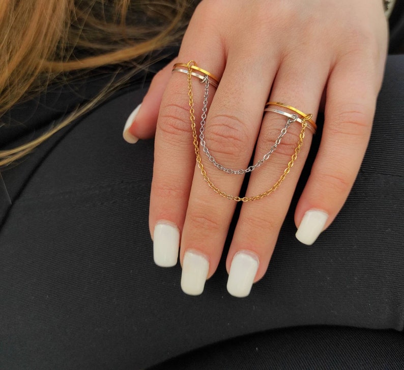 May include: A gold and silver chain ring set. The ring set features two rings connected by delicate chains. One ring is gold, the other is silver. The rings are worn on adjacent fingers. The background is a black fabric.