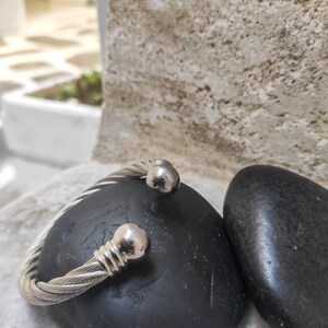 K&ouml;nnte beinhalten: Ein silberfarbenes, geflochtenes Kabel-Armband mit kugelf&ouml;rmigen Endkappen. Das Armband liegt auf einem schwarzen, glatten Stein. Der Hintergrund zeigt eine strukturierte, hellfarbige Steinmauer.