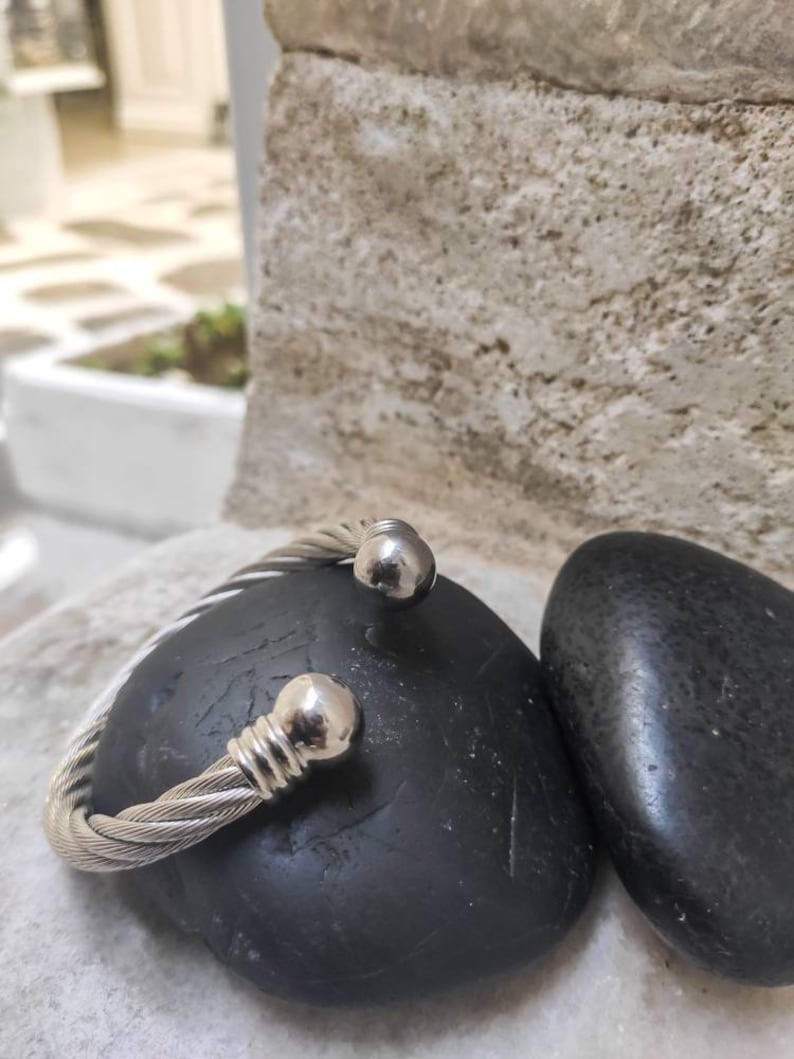 K&ouml;nnte beinhalten: Ein silberfarbenes, geflochtenes Kabel-Armband mit kugelf&ouml;rmigen Endkappen. Das Armband liegt auf einem schwarzen, glatten Stein. Der Hintergrund zeigt eine strukturierte, hellfarbige Steinmauer.
