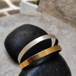 Pode incluir: Duas pulseiras minimalistas, uma prateada e outra dourada, repousam sobre uma pedra preta lisa. As pulseiras s&atilde;o bandas simples, mostrando um design elegante e moderno. A pulseira dourada tem um acabamento polido, enquanto a prateada &eacute; mate.