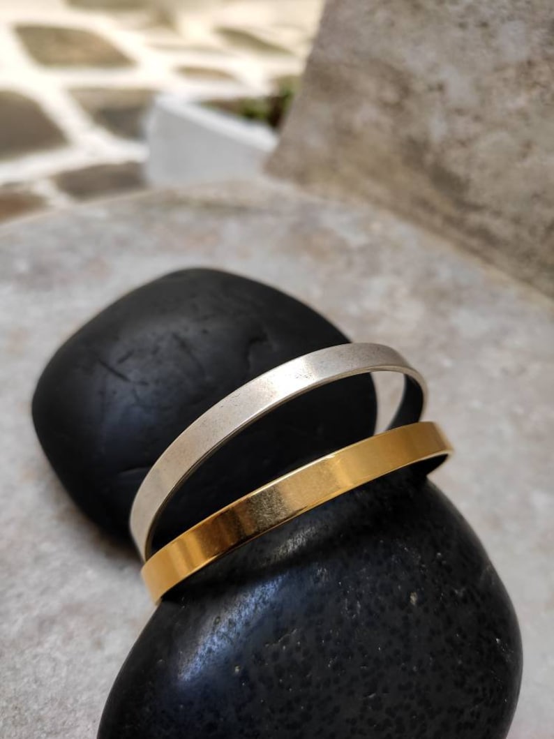 Pode incluir: Duas pulseiras minimalistas, uma prateada e outra dourada, repousam sobre uma pedra preta lisa. As pulseiras s&atilde;o bandas simples, mostrando um design elegante e moderno. A pulseira dourada tem um acabamento polido, enquanto a prateada &eacute; mate.