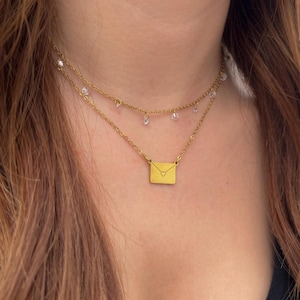Puede incluir: Collar dorado en capas con un pequeño colgante de sobre y cristales transparentes colgantes. El collar se lleva alrededor del cuello, una delicada pieza de joyería.