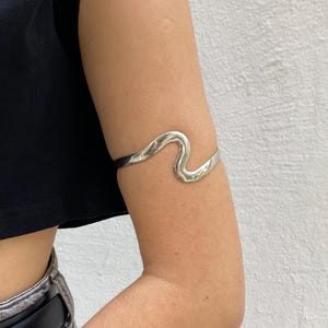 May include: A silver arm cuff with a wavy design. The cuff is worn on the upper arm and has a polished finish. The jewelry is a modern accessory.