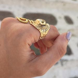 May include: A gold ring with an elaborate design, featuring a large, rounded stone. The ring has a textured band and a decorative swirl element. The ring is worn on a finger.