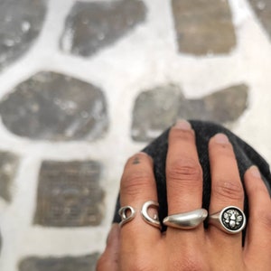 Op de afbeelding: Een hand met verschillende zilveren ringen. De ringen hebben ontwerpen zoals een wervel, een halve maan, een gladde band en een zegelring met een bijenmotief. De achtergrond is een stenen oppervlak.