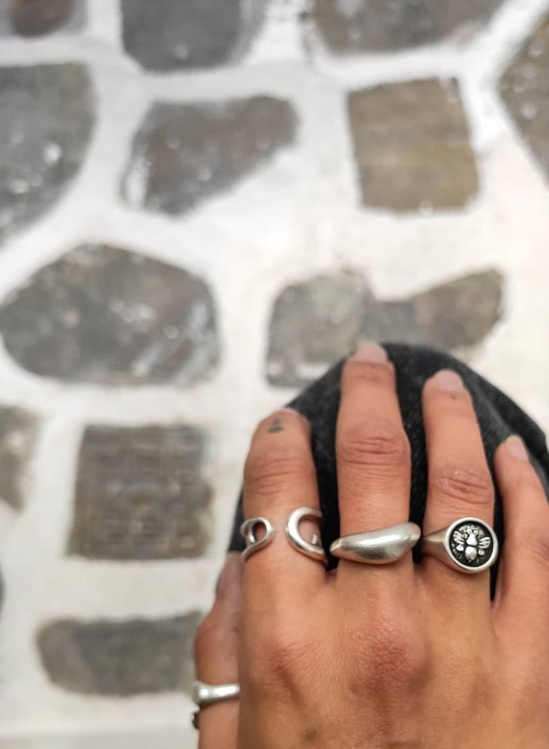 Op de afbeelding: Een hand met verschillende zilveren ringen. De ringen hebben ontwerpen zoals een wervel, een halve maan, een gladde band en een zegelring met een bijenmotief. De achtergrond is een stenen oppervlak.