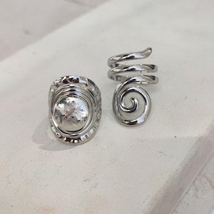 May include: Two silver rings. One ring has a textured, circular design. The other ring is a spiral.