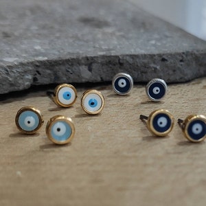 Puede incluir: Pendientes de botón con diseño de ojo. Los pendientes tienen un engaste dorado o plateado con un detalle de ojo azul y blanco. Se muestran sobre una superficie de madera.